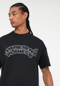 Melna kokvilnas T-kreklu ar īsām piedurknēm, uz kura ir drosmīgs, izliekts grafisks teksts "ŠEIT NAV PLĀNÁ B" pelēkā krāsā. Vienkāršs apaļais kakla izgriezums.