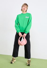 Sudadera verde con logo, pantalones anchos negros y zapatillas beige. Sosteniendo un bolso suave rosa. Fondo liso con suelo verde.