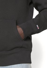 Felpa nera con polsini e orlo a costine, dotata di tasca frontale e di una piccola patch con logo nei colori rosso, bianco e blu sulla manica.