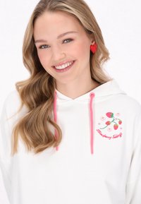 Felpa bianca con coulisse rosa, caratterizzata da fragole e fiori ricamati. Tessuto morbido con una vestibilità ampia. Orecchini rossi a forma di cuore.