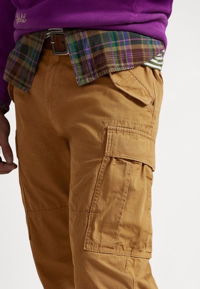 Béžové cargo nohavice s viacerými bočnými vreckami, vyrobené z odolnej látky. Kombinované s károvanou košeľou pod fialovou mikinou.
