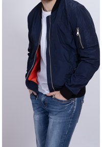 Námořnická bombarďáková bunda s zipovým uzávěrem, žebrovanými manžetami a oranžovou podšívkou. Obsahuje kapsu na zip na rukávu a má hladkou texturu.