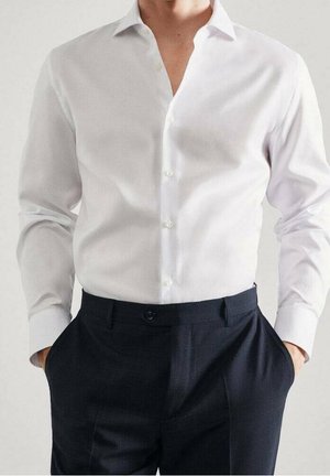 Chemise blanche à boutons, avec un col décontracté, associée à un pantalon à carreaux foncés. Le tissu est lisse, présentant une coupe élégante et ajustée.