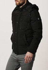 Czarna pikowana kurtka z wysokim kołnierzem i kapturem, z przednim zamkiem błyskawicznym i zatrzaskami, kieszenią na lewej piersi oraz logo na rękawie.
