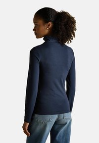 Kihara naiste juustega naine, kellel on kuklas kõrgel kinni pandud juuksed, kannab tumesinist pika varrukaga toppi, millel on laineline kõrge kaelus, ja siniseid teksaseid, vaadates vasakule.