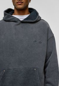 Grijze oversized hoodie met een getextureerde fleece voorzak, capuchonvoering in een donkerder grijs en een subtiel geborduurd logo op de borst.