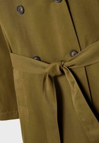 Trench coat verde oliva con una cintura di tessuto legata e bottoni scuri visibili sul davanti.