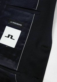 Granatowa marynarka o gładkiej tkaninie i teksturowanym splocie, z białą metką z czarnym logo, kontrastowym przeszyciem i dopasowanymi szczegółami.