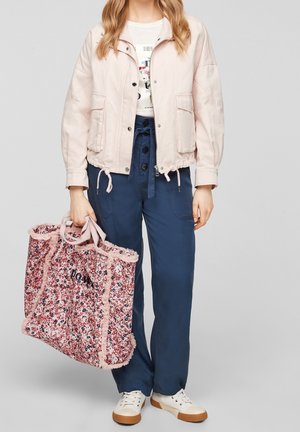 Femme portant une veste rose clair et un pantalon bleu, tenant un grand sac fourre-tout à motif floral et portant des baskets blanches.