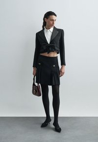 Czarna skórzana marynarka z kokardą, biała koszula z kołnierzykiem, czarna spódnica plisowana, czarne rajstopy, czarne buty, trzymając brązową torebkę.