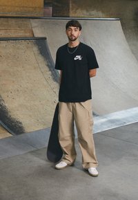 Svart Nike SB t-shirt, beige cargo byxor och vita sneakers; en skateboard under armen. Inomhusskatepark med träramper i bakgrunden.