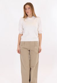 Jeune femme aux longs cheveux blonds portant un pull blanc à manches courtes et un pantalon beige à jambes larges, debout devant un fond blanc.
