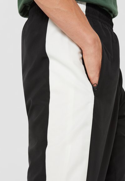 Pantalons de survêtement noir et blanc avec une poche latérale zippée, présentant un panneau texturé blanc et une finition en tissu noir lisse. Taille élastique.