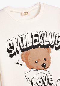 Sudadera blanca con cuello redondo, con un gráfico de un oso de peluche marrón y el texto negro "SMILECLUB LOVE", con acentos de estrellas. Textura de tela suave.
