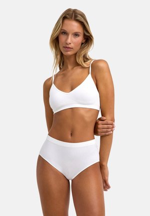 Kobieta o blond włosach, ubrana w biały bralet i białą bieliznę z wysokim stanem, stojąca na jasnym, jednolitym tle.