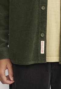 Dunkelgrünes Cordhemd mit Knopfverschluss, das ein gesticktes Etikett mit der Aufschrift "REVOLUTION" trägt. Darunter wird es über einem hellbeigen Oberteil und schwarzen Hosen getragen.