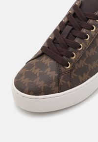 Gros plan d'une basket marron avec des lacets marron foncé, des œillets dorés et une surface texturée présentant un motif répété du logo "MK" sur une semelle blanche.