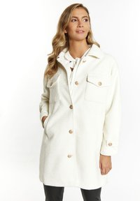 Witte textuur coat met grote knopen, twee borstzakken en een kraag. Het materiaal lijkt zacht en warm, geschikt voor het dragen in lagen.