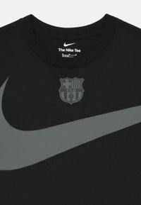 Μαύρο μπλουζάκι Nike με στρογγυλή λαιμόκοψη, διακοσμημένο με ένα μεγάλο γκρι λογότυπο Nike και ένα γκρι έμβλημα της FC Barcelona τυπωμένο μπροστά.