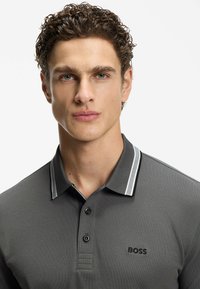 Szara koszulka polo z teksturowanej tkaniny, z dwoma czarnymi guzikami i kołnierzem w kontrastowe paski. Na piersi znajduje się czarne logo "BOSS".