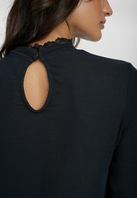 Mörk ribbad topp med design som har en nyckelhålsöppning bak, utsmyckad med spetskant och knappstängning. Tyget ser mjukt och texturerat ut.
