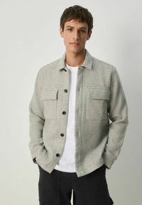 Grau kariertes Hemd mit zwei Brusttaschen, Knopfverschluss und weichem Stoff-Texture, über einem weißen T-Shirt getragen. Schwarze Hose.