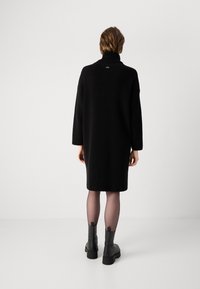Zwarte gebreide jurk met hoge hals, knie-lange snit en losse silhouet. Gecombineerd met zwarte enkellaarsjes en doorschijnende tights.