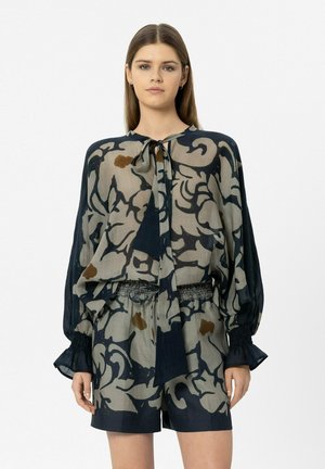 Junge Frau trägt eine graue und schwarze gemusterte Bluse mit Schlips und passende lockere Shorts, steht vor weißem Hintergrund.