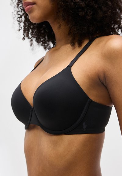 Γυναίκα που φοράει μαύρο επενδυμένο σουτιέν με λεπτές τιράντες και μπανέλα, δείχνοντας το πάνω μέρος του σώματος και τα σγουρά μαλλιά της.
