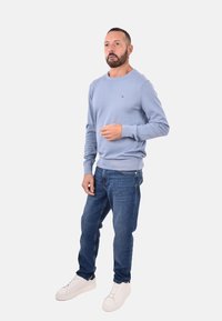 Uomo con barba e orecchino in piedi indossando un maglione azzurro chiaro, jeans blu e sneakers bianche contro uno sfondo chiaro e semplice.