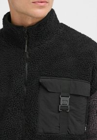 Chaqueta de forro polar negra con cuello alto, con una superficie texturizada y un bolsillo frontal con una correa y detalle de hebilla. Cierre de cremallera visible.