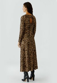 Vrouw in een lange jurk met luipaardprint, voorzien van een sleutelgatopening aan de achterkant en een strikdetail, gecombineerd met zwarte haklaarzen, staand tegen een effen achtergrond.