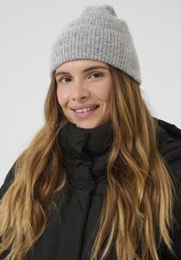 Kvinde med langt lystbrunt hår, der smiler, iført en grå strikket hue og en sort vinterjakke med høj krave mod en ensfarvet baggrund.