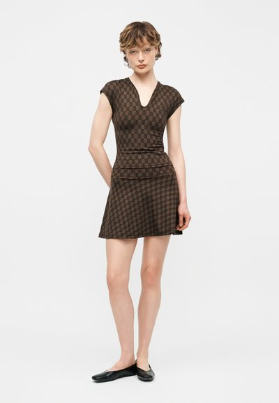 Γυναίκα που φοράει εφαρμοστό καφέ φόρεμα με σχέδιο, κοντά μανίκια, V-λαιμόκοψη και μαύρα παπούτσια φλατ, στέκεται μπροστά σε απλό λευκό φόντο.