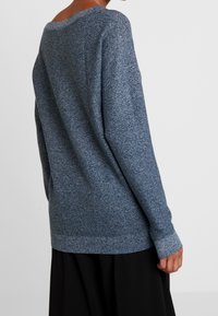 Pull en maille bleu foncé avec une finition texturée, doté d'un large encolure et de manches longues, porté sur un vêtement noir.