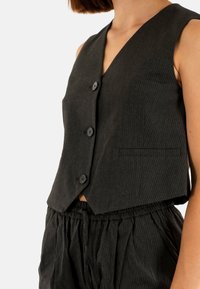 Gilet nero a righe sottili con scollatura a V, dotato di tre bottoni e una piccola tasca laterale. Il tessuto appare strutturato e leggero.