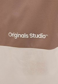 Textile marron et blanc avec un motif en grille ; comporte le texte "Originals Studio™" en lettres blanches, positionné sur la section marron.
