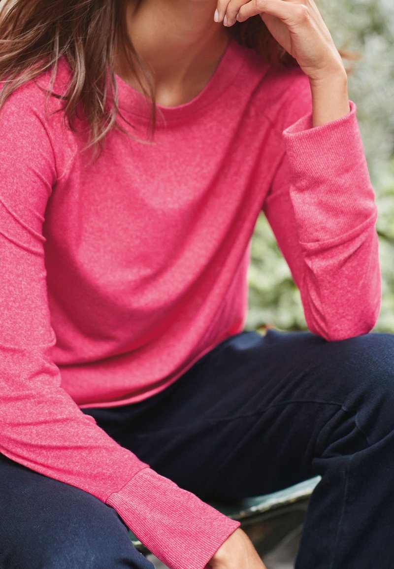 Haut en maille rose à manches longues avec un col rond et des poignets côtelés, associé à un pantalon sombre, sur un fond vert flou.