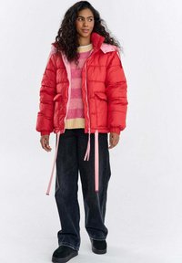 Czerwona kurtka puchowa z różowym kołnierzem i suwakiem, z kieszeniami i ozdobnymi wstążkami, noszona na różowym swetrze w paski i ciemnych spodniach.