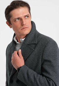 Grå uld peacoat med høj krave og dobbeltradede knapper, båret over en lysegrå sweater, fremhævet af et armbåndsur.