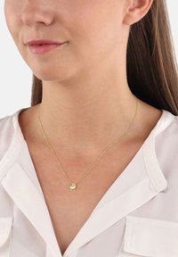 Kobieta nosząca cienki złoty łańcuszek z małym, gładkim wisiorem w kształcie serca na białej bluzce.