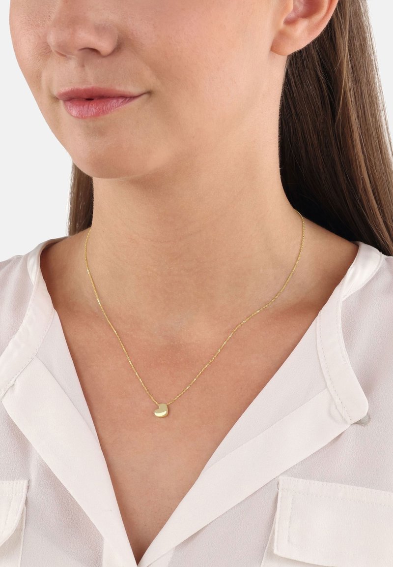 Kobieta nosząca cienki złoty łańcuszek z małym, gładkim wisiorem w kształcie serca na białej bluzce.