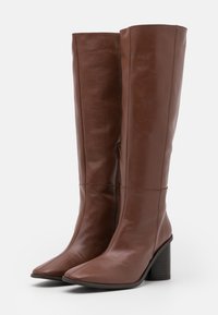 Botas de piel marrón hasta la rodilla con puntera cuadrada, tacón ancho, cremallera lateral y textura suave. Diseñadas para una silueta ajustada.