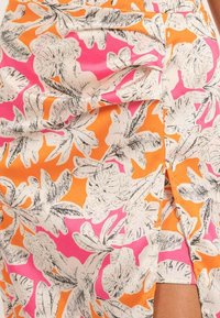 Jupe enroulée florale dans des tons vifs d'orange, de rose et de noir. Présente une haute fente et un tissu texturé avec un motif de feuilles tropicales.