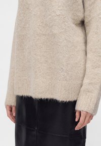 Pull en maille beige duveteuse à manches longues associé à une jupe en cuir noire, mains détendues sur les côtés sur un fond neutre.