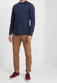 Man draagt een navy blauwe knopenhemd, tan opgerolde chino's en bordeauxrode sneakers, staande tegen een effen witte achtergrond.