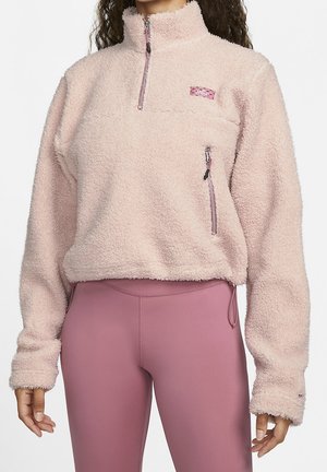 Femme portant un pull polaire rose clair avec des poches à fermeture éclair et des leggings mauve taille haute, vue du cou aux hanches, mains détendues.