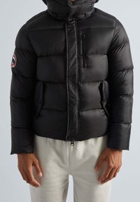 Veste matelassée noire avec un col haut, une poche poitrine zippée et des poches latérales ; caractérisée par une texture matelassée et des poignets élastiques.