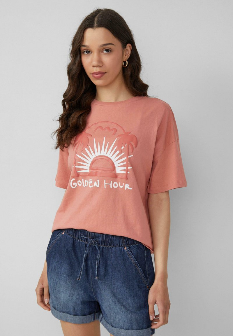 T-shirt en coton rose avec une coupe décontractée, ornée d'un graphique blanc représentant un soleil et des palmiers. Porté avec un short en denim bleu.