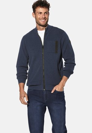 Marineblauer Reißverschluss-Pullover aus strukturiertem Stoff, mit einer Reißverschlusstasche und gerippten Bündchen und Saum. Kombiniert mit dunklen Jeans.